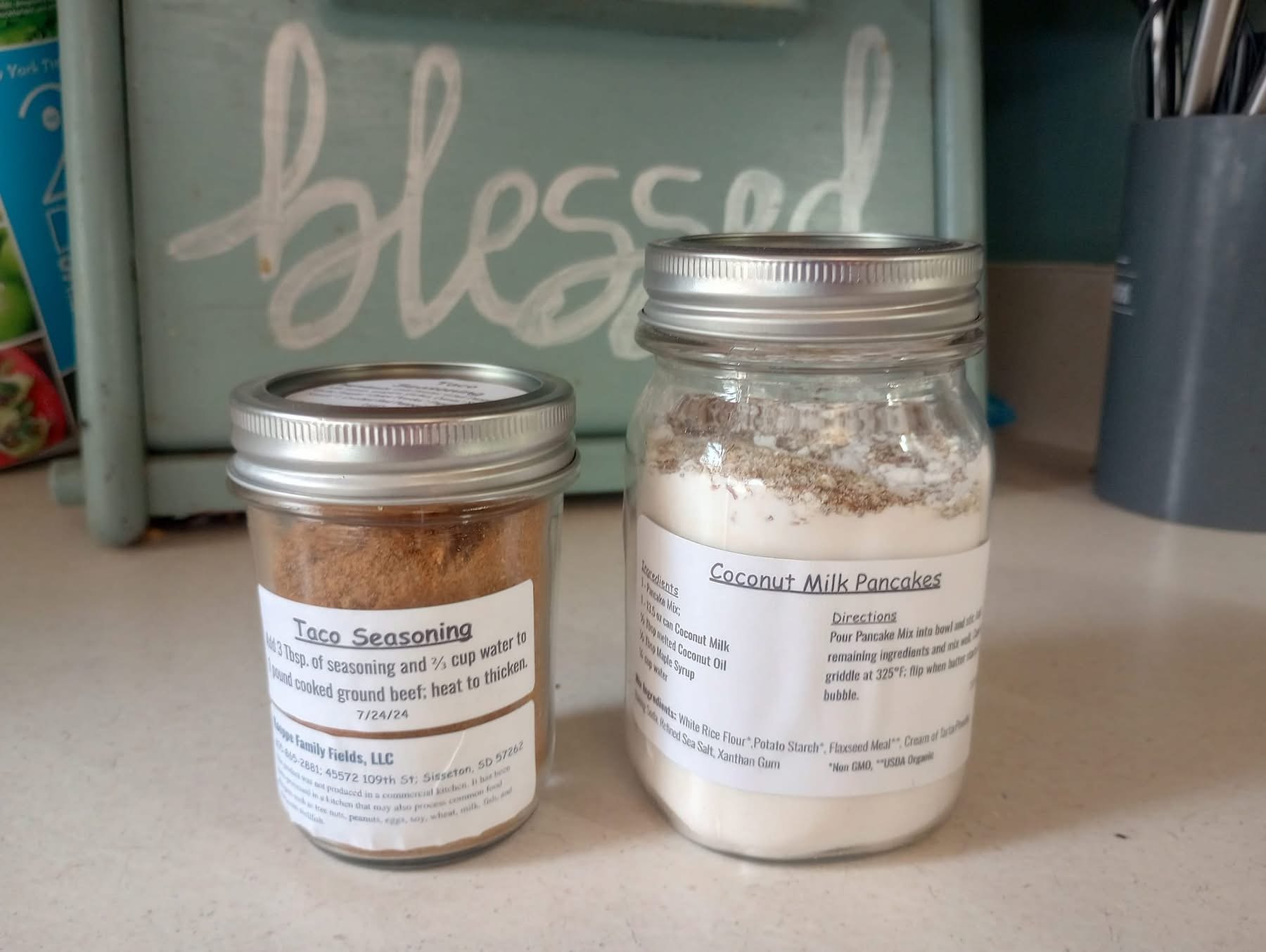 Glass jars containing taco seasoning and coconut milk pancake mix on a kitchen countertop, with a decorative "blessed" sign in the background. The taco seasoning jar has usage instructions, while the coconut milk pancake jar lists ingredients and directions for preparation.