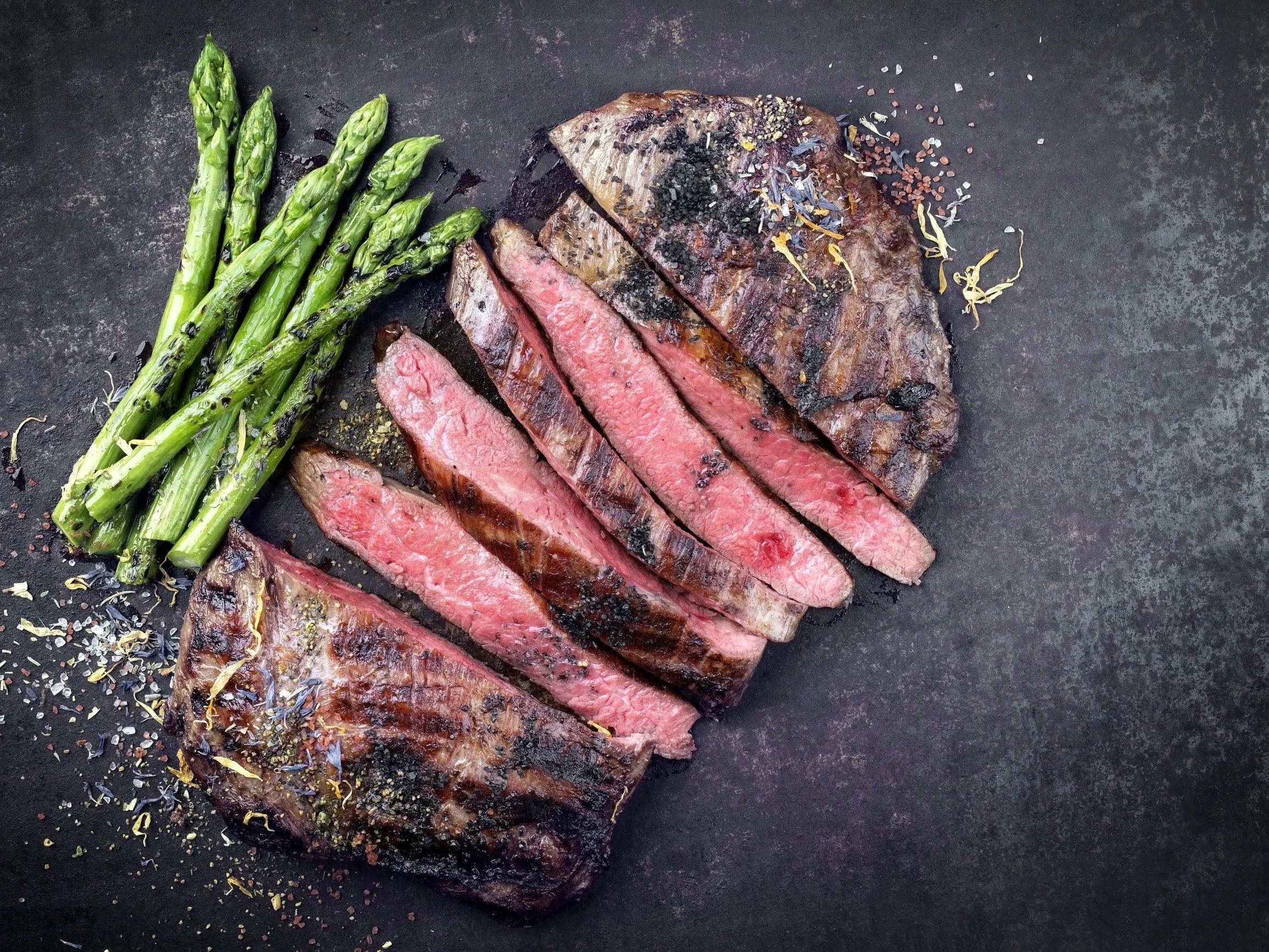 Grilled steak slices served with charred asparagus on a dark textured surface, garnished with various spices and herbs. Perfect for showcasing a delicious meal or barbecue dish.